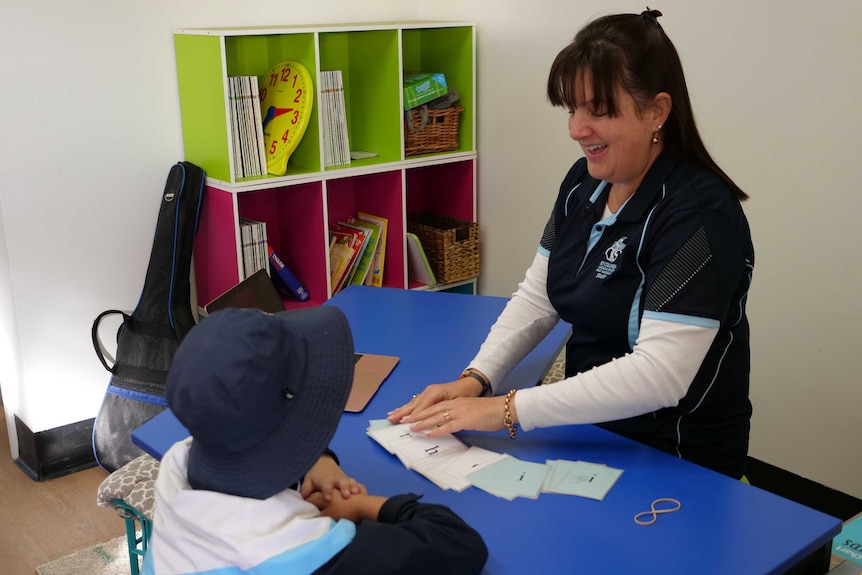 A female teacher sits at a desk with a young male student, showing cards with letters on them.