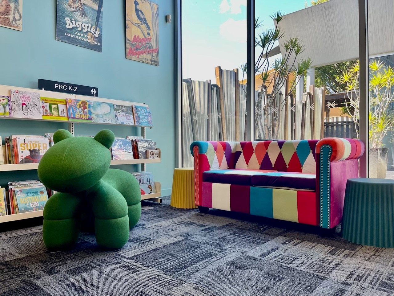 A kids' section at a library with a large green stuffed dinosaur, a colourful couch and books on shelves