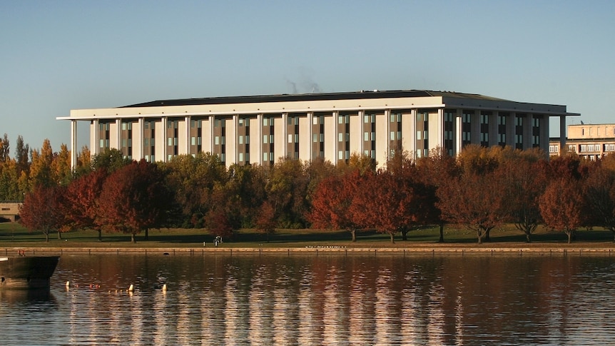 A neo-classical building surrounded by trees by a lake shore.