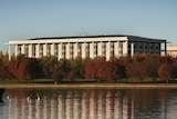 A neo-classical building surrounded by trees by a lake shore.