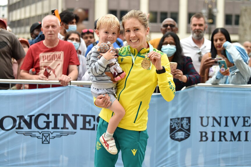 An Australian athlete smiles at the camera holding a little boy and her gold medal. 