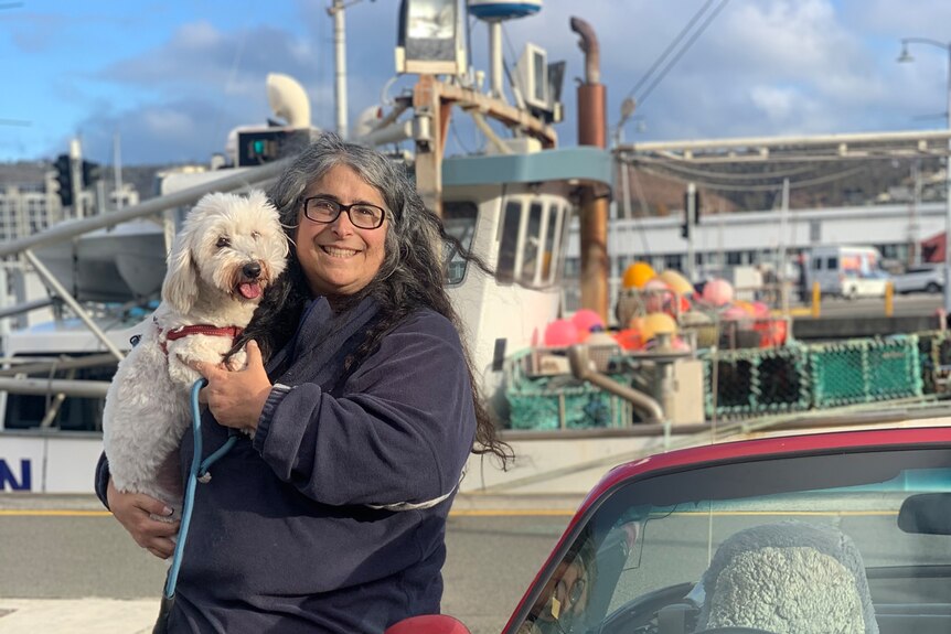 A woman with long dark hair and dark-framed spectacles poses for a photo on a harbour, next to a red car, holding her small dog