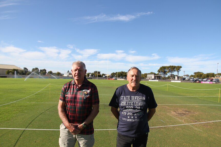 Two older man stand on an oval in front of a cricket pitch beneath a sunny sky.