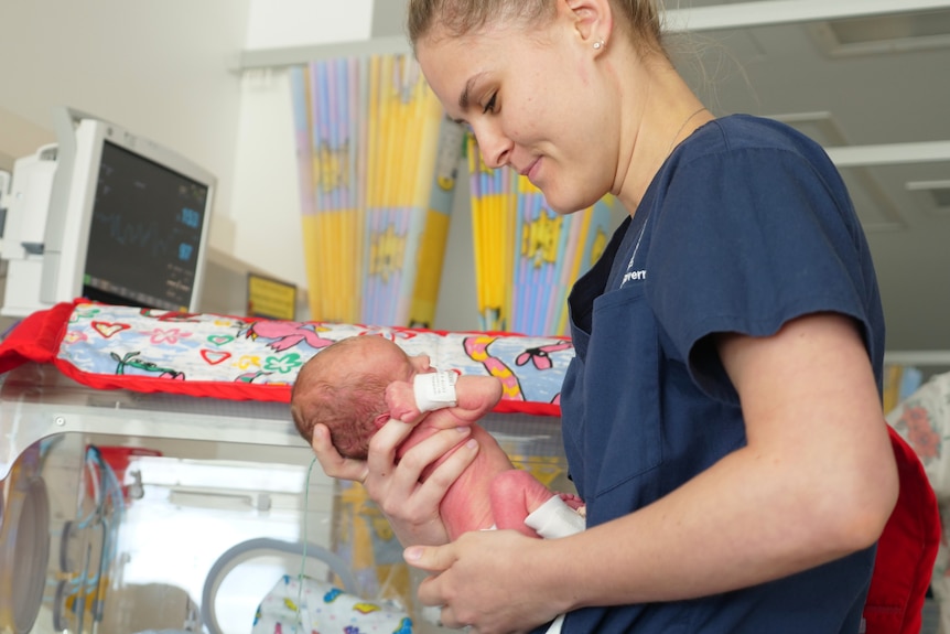 Charlotte smiling at baby Kove who she is holding in her hands, incubator behind.