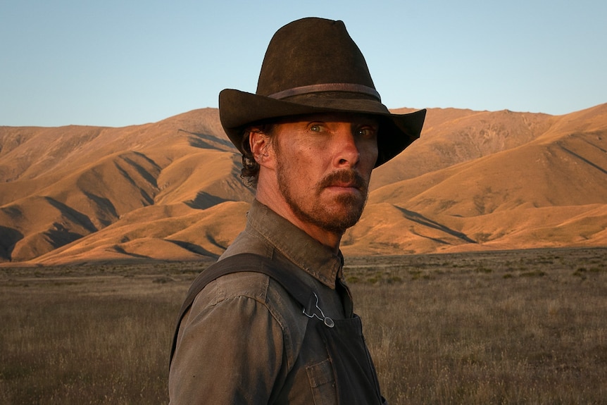 A scruffy 40-something man in a cowboy hat stands in a field, ringed by table hills, looking intensely into the camera lens