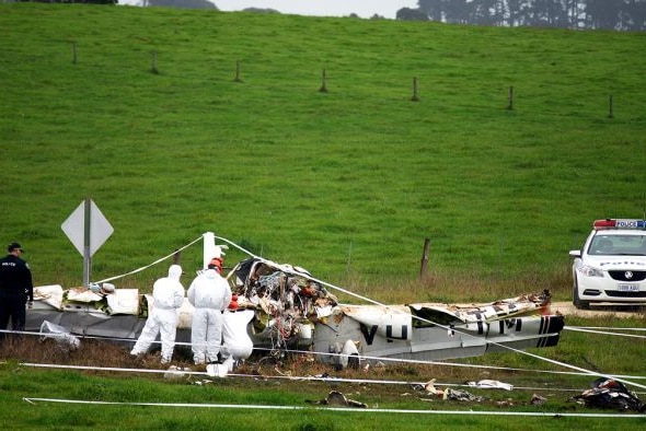 A wrecked plane sits in the middle of a green pasture and hill, forensic investigators in white suits and police attend the scen