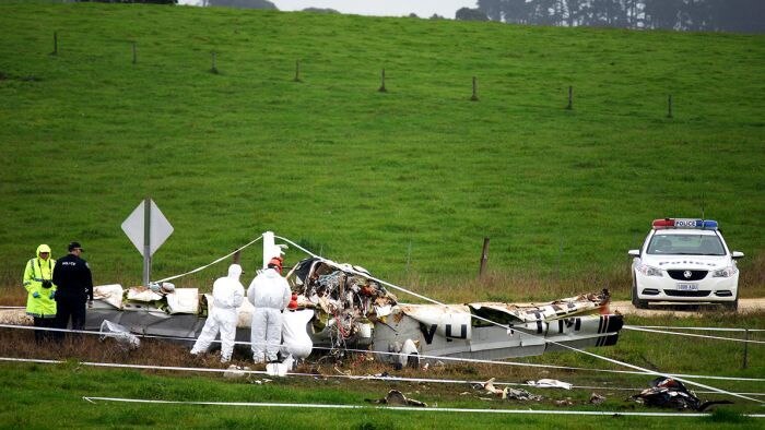 A wrecked plane sits in the middle of a green pasture and hill, forensic investigators in white suits and police attend the scen