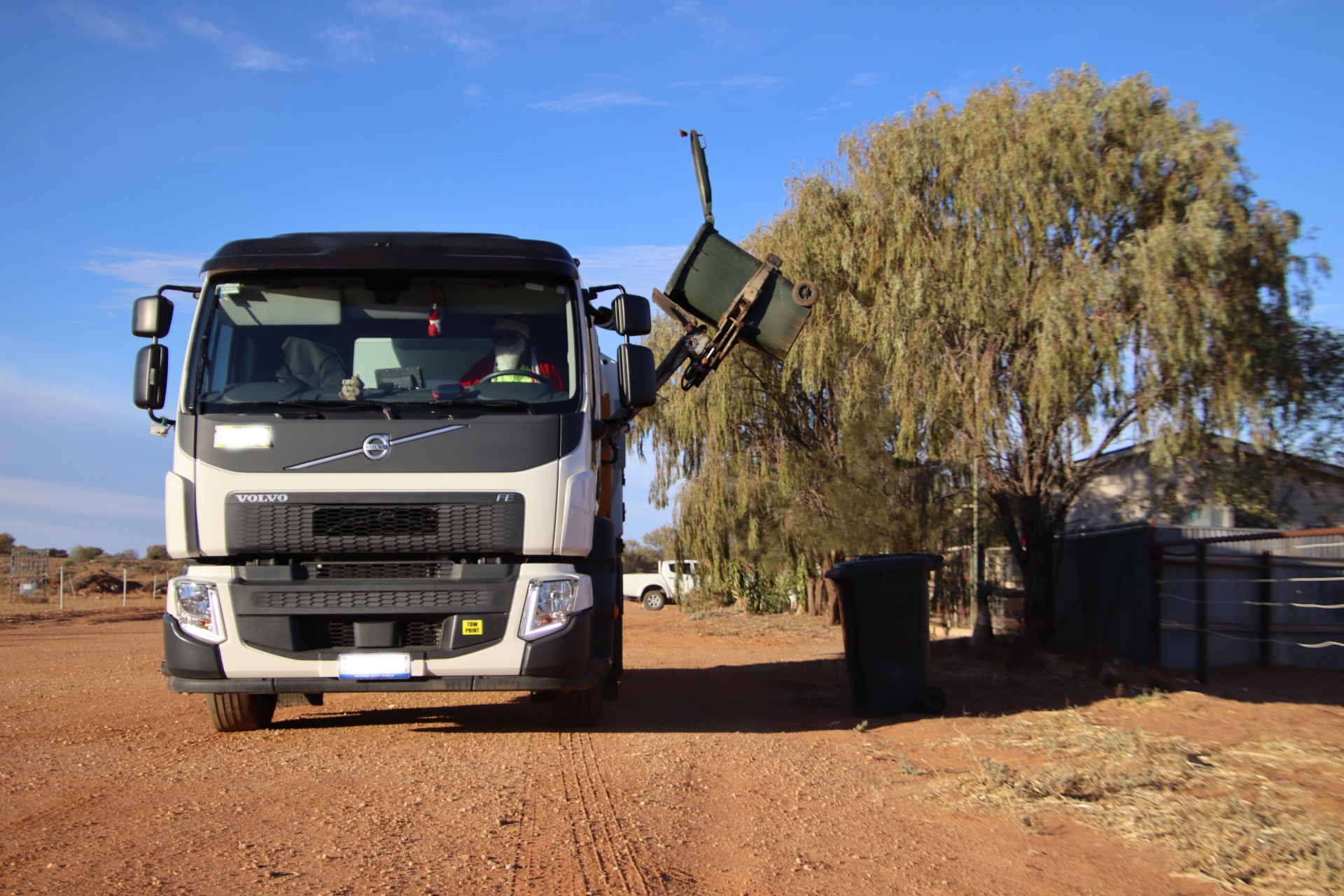 Un camión de basura blanco levanta un bote de basura en un camino de tierra al lado de una casa.