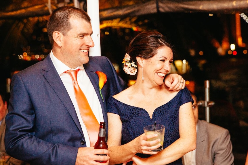 A man and a woman smile big on their wedding day, both wearing navy blue and holding a drink at the reception
