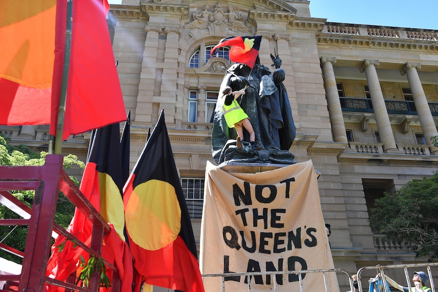 A protestor with an Aboriginal flag on a statue and a sign "Not the Queen's land".