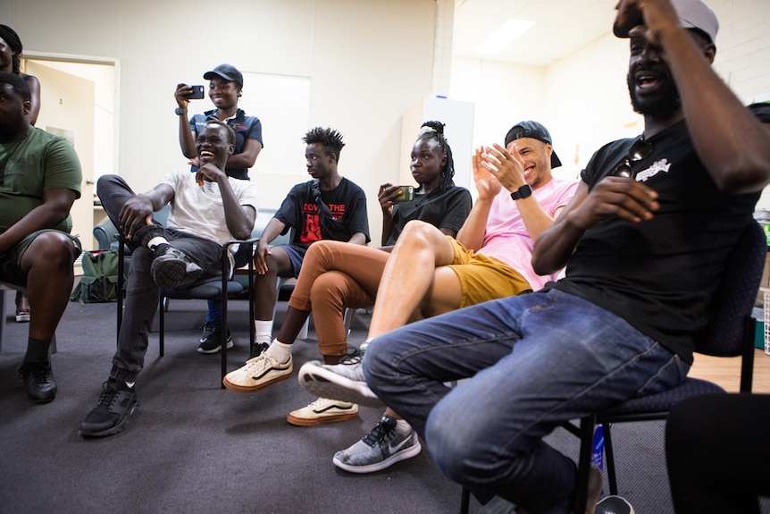Young men and women sit on plastic chairs with cast members from Barbershop Chronicles, mid conversation and laughing.