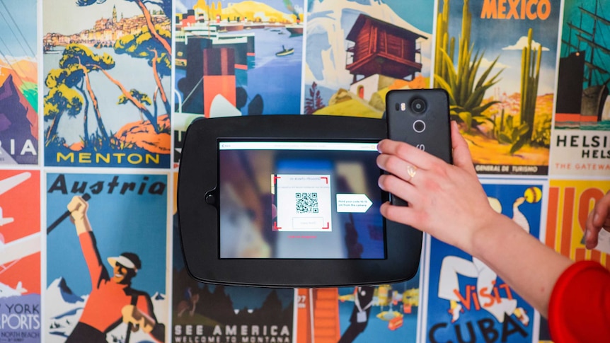 A photo of a hand holding a mobile phone scanning a QR code at a kiosk with travel posters on the wall behind