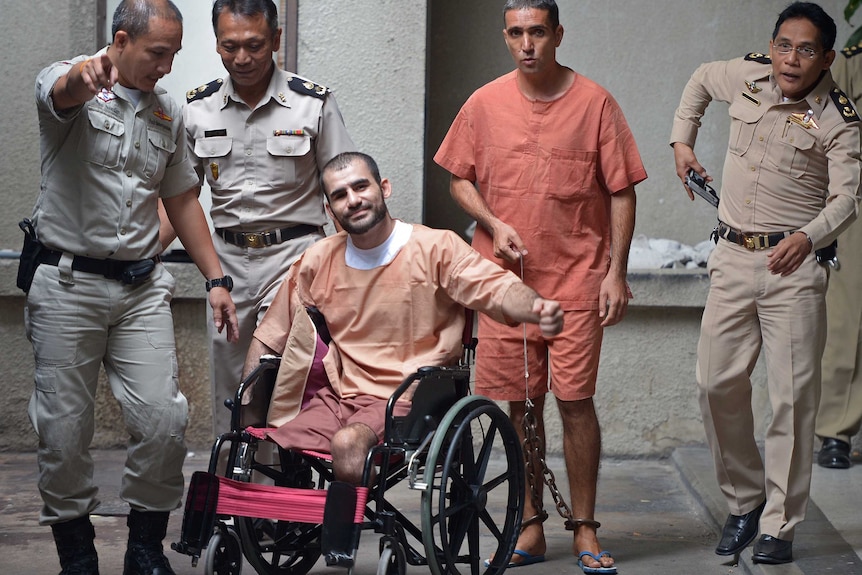 Thai police flank a man in a wheelchair in orange prison uniform next to a fellow prisoner walking beside him.