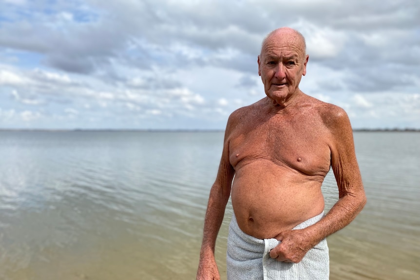 An older man stands in water. He has a white towel around his waist and no shirt. he has tanned skin and some wrinkles. He frown
