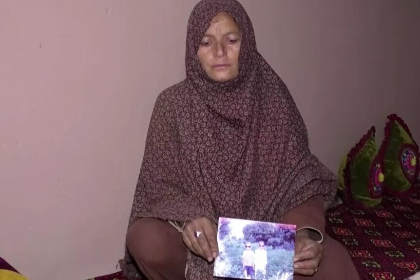 Pakistani woman with hijab sitting on floor, solemn expression, holding a photo of her two sons.