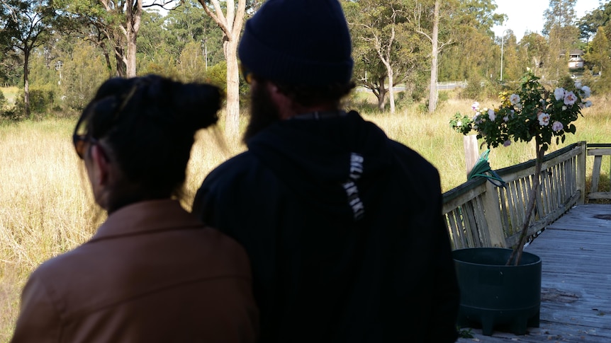 A man and woman stand silhouetted on an old veranda looking out at sunny trees and grass, wearing beanie and sunglasses.