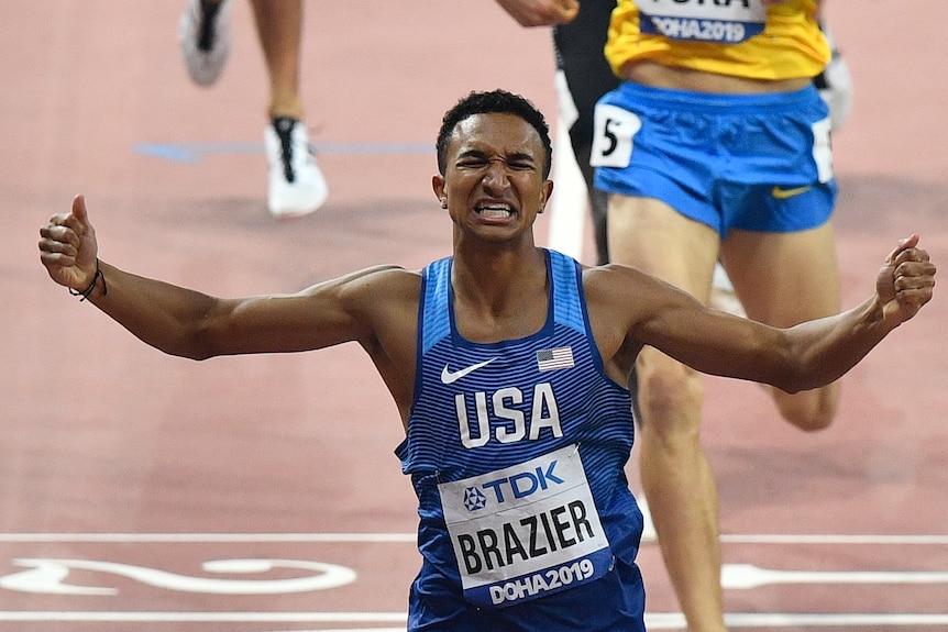 Donavan Brazier runs with his eyes scrunched up and mouth in a grimace, both arms in the air wearing a blue singlet with USA on