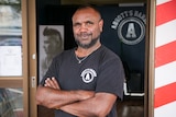 An Aboriginal man standing out the front of his barber shop with his arms crossed a smile on his face.