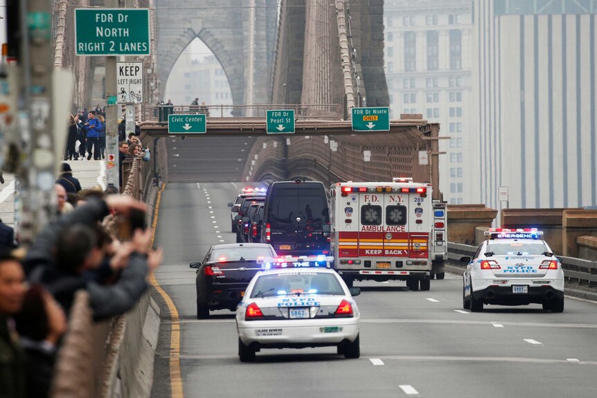 People watch and take photographs as police cars and an ambulance vehicle cross an otherwise empty bridge.