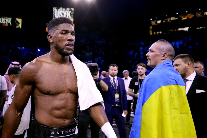 Anthony Joshua walks past Oleksandr Usyk, who is looking at him out of the corner of his eye while draped in a Ukraine flag
