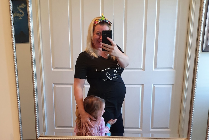 A pregnant woman takes a selfie in a mirror with a little girl hugging her