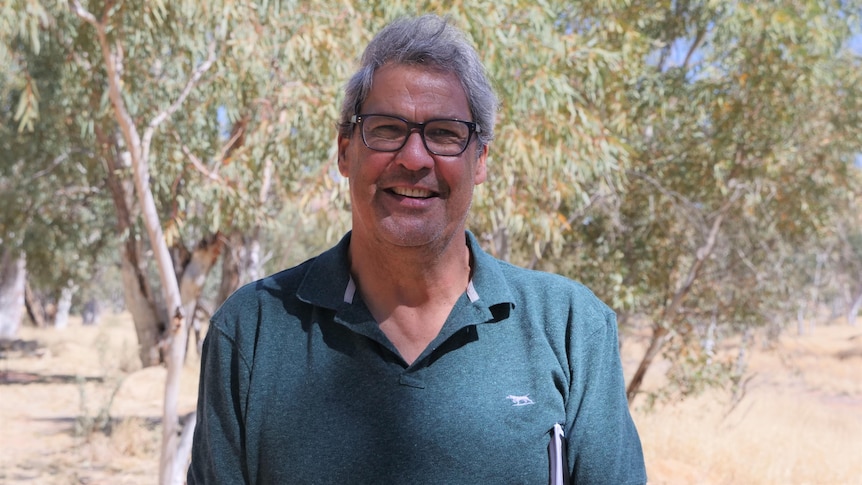 man wearing glasses and green shirt standing in centre of frame, smiling, in front of bushland