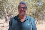 man wearing glasses and green shirt standing in centre of frame, smiling, in front of bushland