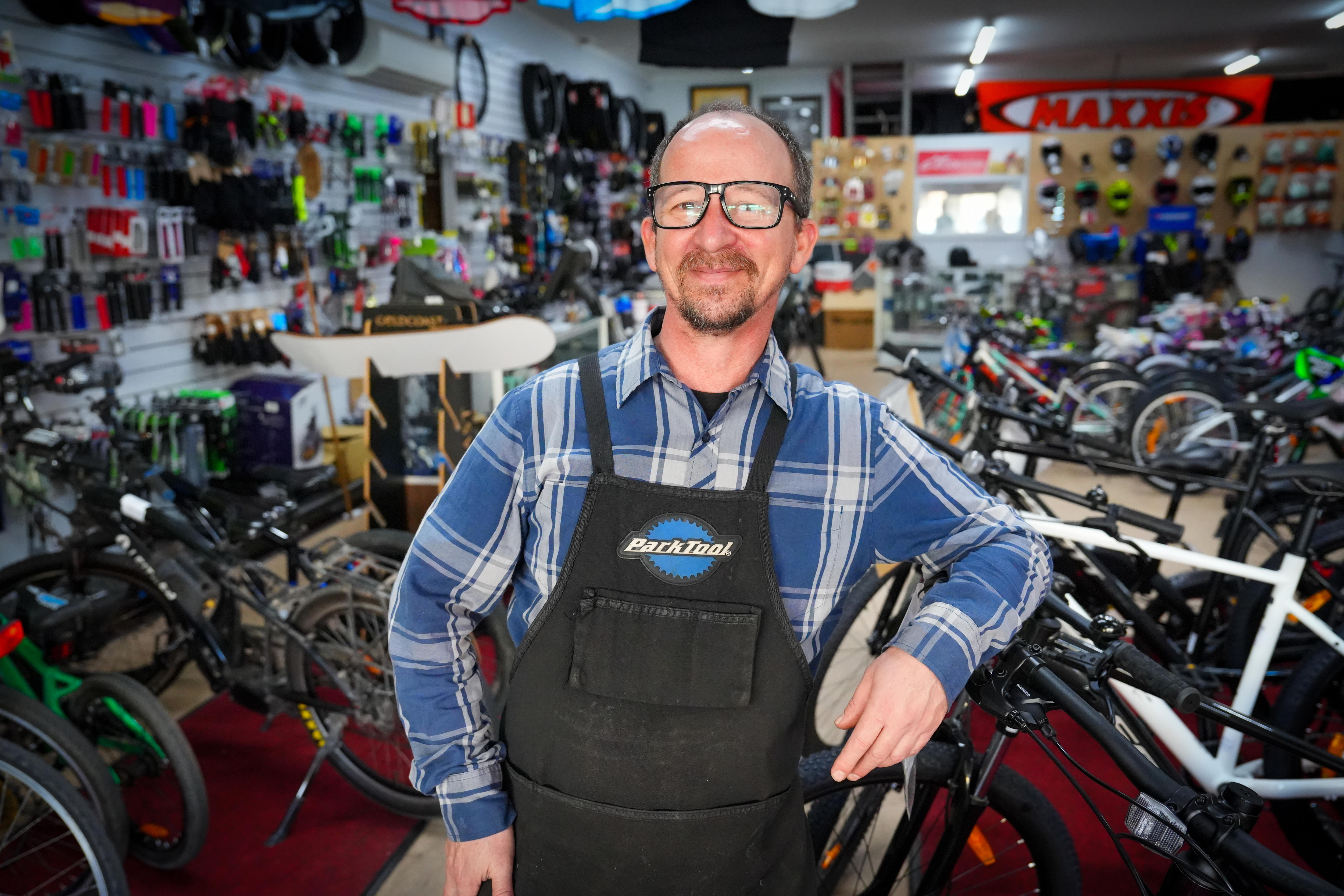 A man in a blue shirt and dungarees standing inside a bicycle shop.
