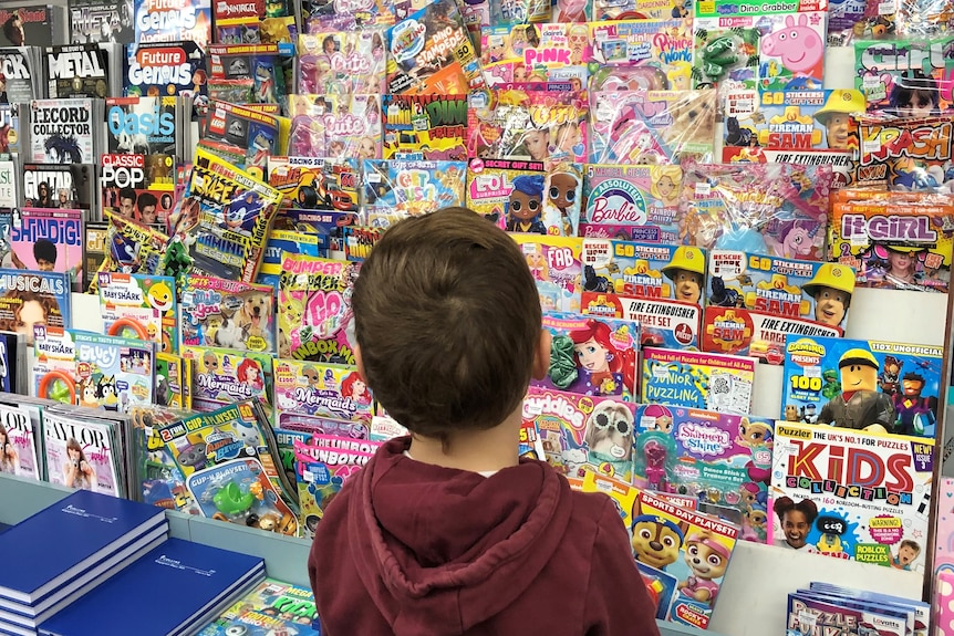 young boy stand with his back to the camera, looking at a stand of kids magazines in a newsagency