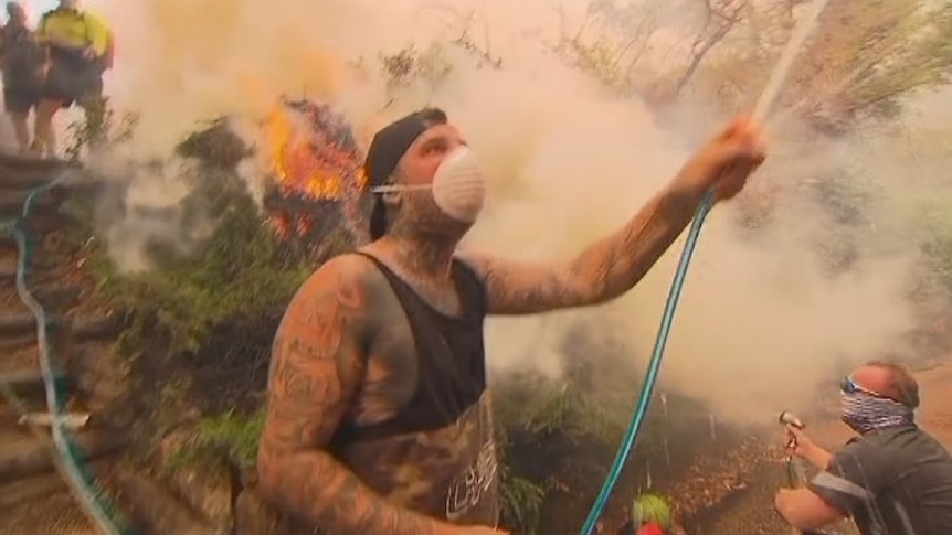 A man wearing a disposable dust mask hoses down a house in Menai in southern Sydney to defend it from bushfire.