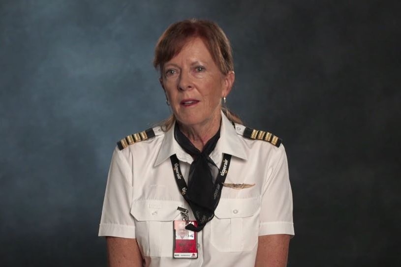 A woman in pilot uniform with Tigerair lanyard sits before grey backdrop, with neutral expression.