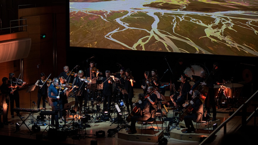 The Australian Chamber Orchestra perform on a dark stage with a video project in the background of a river plain.