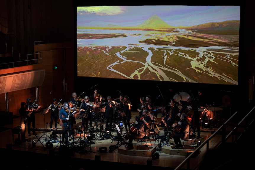 The Australian Chamber Orchestra perform on a dark stage with a video project in the background of a river plain.