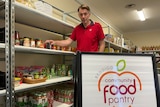 A person standing in pantry, beside some shelving and a sign saying "Bendigo Community Food Pantry"