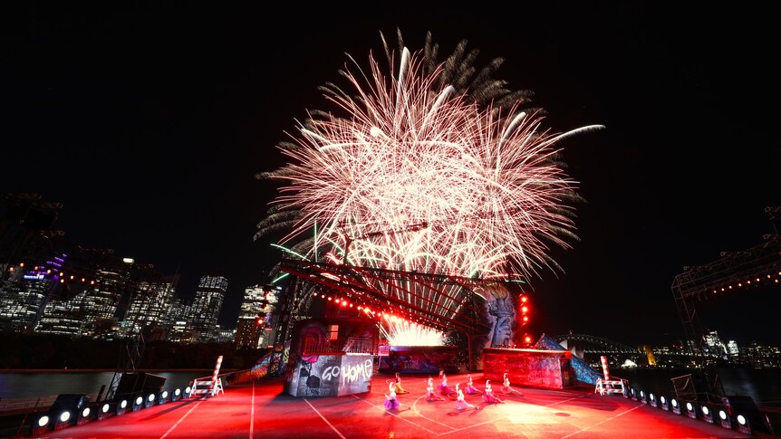 Fireworks explode behind a floating stage with eight dancers performing on a basketball court in an urban setting.
