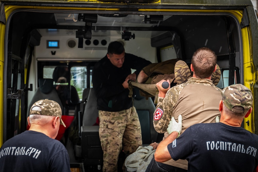 A group of paramedics with camouflage print pants lift a soldier on a stretcher from inside an ambulance van