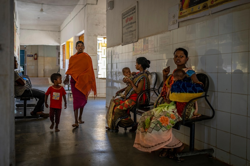 women in colourful dress sit in a hospital hallway with their children.