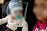 A baby and a young woman in a car. Both have their faces blurred.