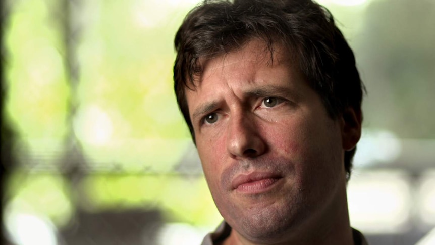 Patrick Dunn, wearing grey shirt, with brown hair, stares sideways pensively, with blurred house background.