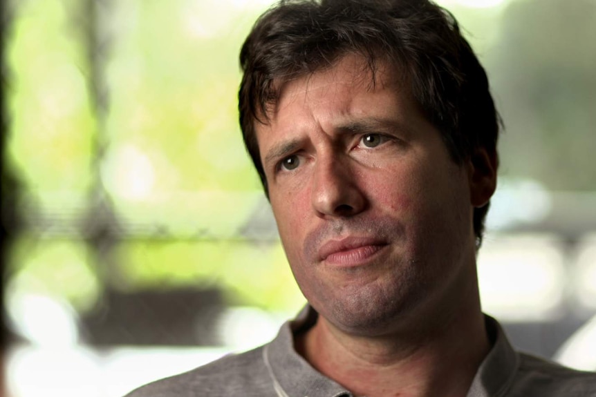 Patrick Dunn, wearing grey shirt, with brown hair, stares sideways pensively, with blurred house background.