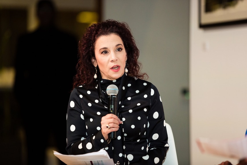 Madeline Di Nonno, dressed in a spotted shirt, holds a mic to her face, she is mid-speech.