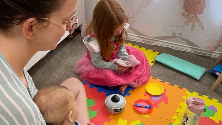 A mother sits with a baby in her lap watching her little girl as she plays with a doll