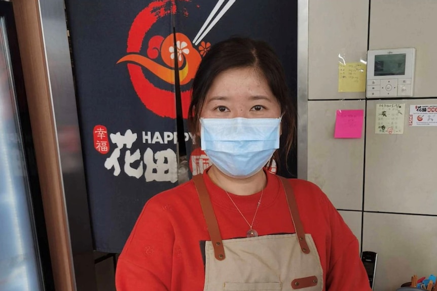Min Ai stands in her restaurant looking at the camera, wearing an apron and surgical face mask. 