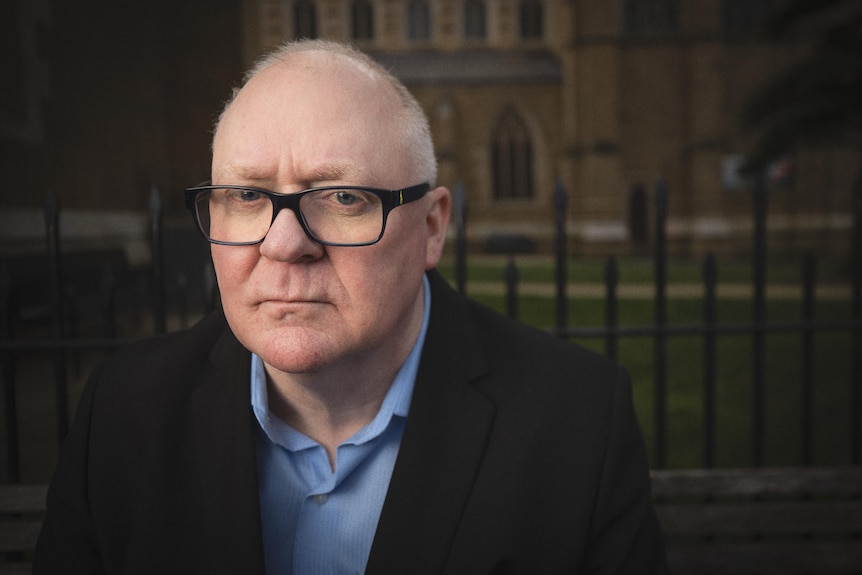 A balding man wearing glasses sits in front of a fence