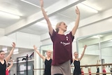 A Queensland Ballet teaching artist in a maroon top and pink skirt leads a class of ballet students in red and black leotards.