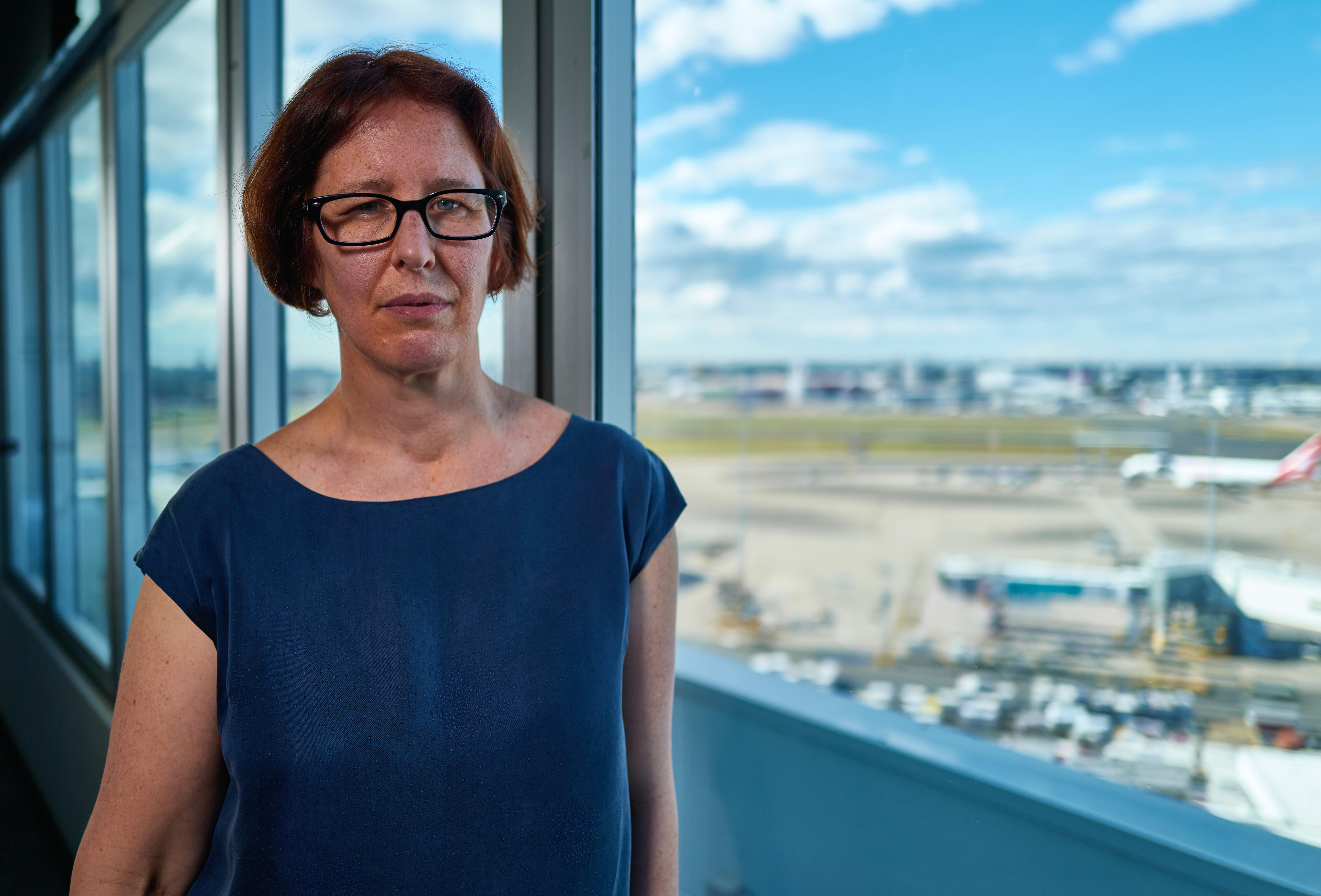 A woman with a neutral expression stands in front of windows that show an airport and planes below.