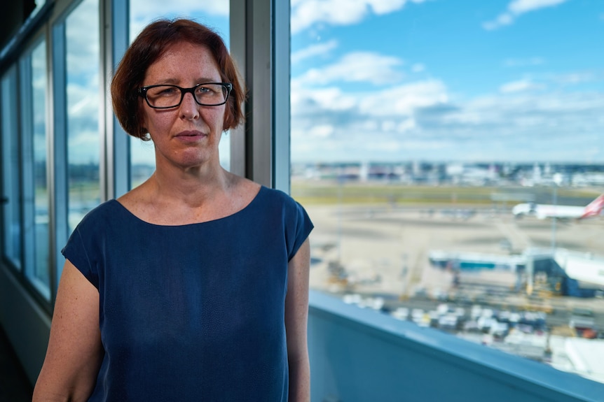 A woman with a neutral expression stands in front of windows that show an airport and planes below.