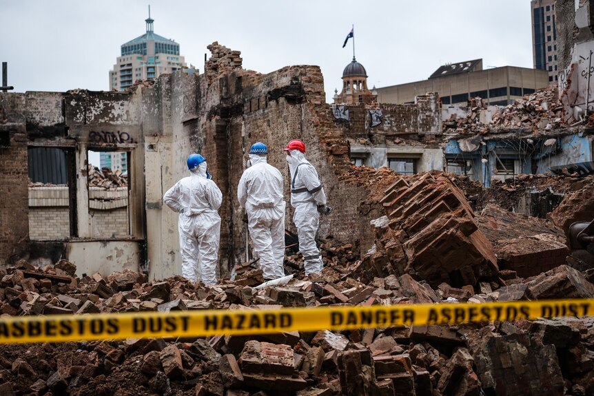 a group of workers wearing white hazmat suits stand in the rubble of a building burnt by fire