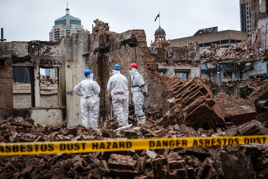 a group of workers wearing white hazmat suits stand in the rubble of a building burnt by fire