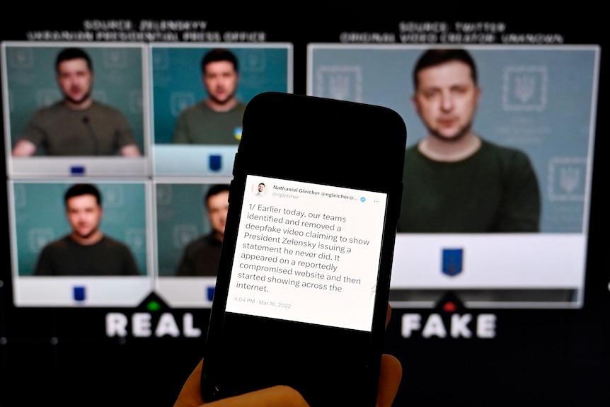 Video screens showing Volodymyr Zelenskyy are behind a phone screen displaying a message about a fake video.